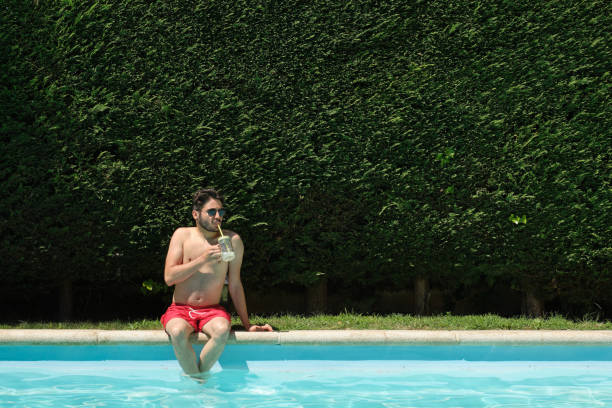 istock Young man wearing sunglasses drinking lemonade sitting on the swimming pool edge. Summer concept. 1328209921