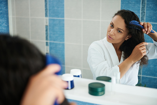 Attractive Hispanic woman wearing white waffle bathrobe looking at her mirror reflection while combing hair in bathroom