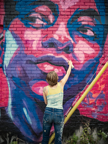 Young woman painting mural using spray paints. She is dressed in casual work clothes. Exterior of uban street, back alley with old brick wall of the house.