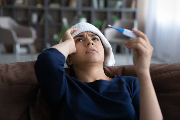 Close up sick Indian woman feeling unhealthy, holding thermometer Close up sick Indian woman feeling unhealthy, holding thermometer, suffering from heat or fever, unhappy young female with towel on head leaning back on couch at home, cold or flu, health problem fever stock pictures, royalty-free photos & images