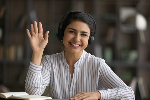 Head shot portrait smiling Indian woman in headphones waving hand and looking at camera, happy businesswoman involved in internet meeting, video call, greeting viewers, teacher leading online lesson