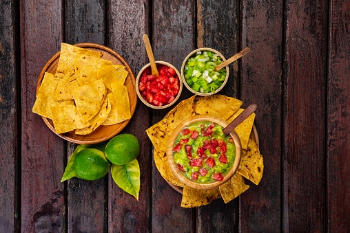 Typical mexican food of guacamole with tortilla chips ,cilantro , tomato, lemon and chives over rustic wooden table in the kitchen.This guacamole the ingredients are avocado, salt, juice of lemon, onion and tomato.