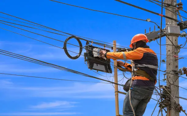 Photo of technician on wooden ladder is installing fiber optic system in internet splitter box on electric pole against blue sky