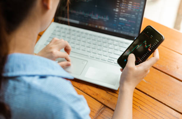 istock Woman's hand with phone and laptop checking stock exchange 1328259304