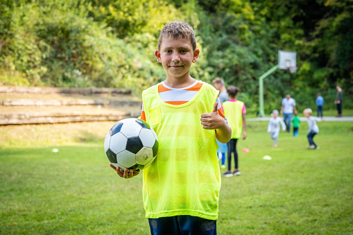 Standing boy with soccer ball, looking at camera and showing thumbs up. Children on football training. Children team sport on fresh air, physical activity and entertainment.