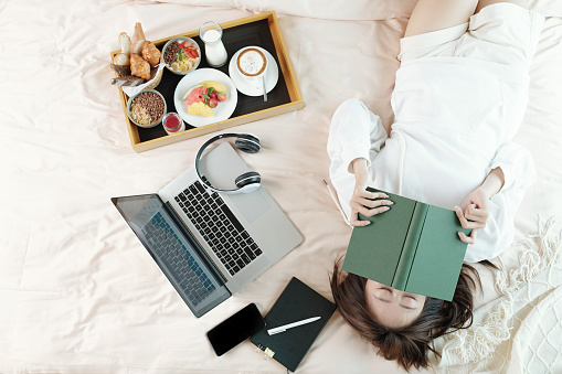 Lazy young woman lying on bed with opened laptop and tray with breakfast, sleeping after reading boring book