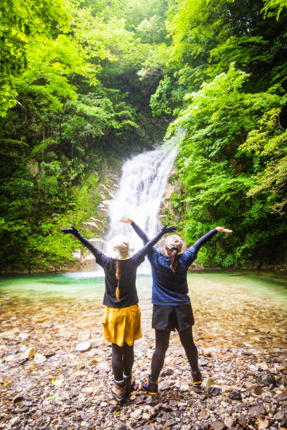 Two Japanese female hikers friends looking towards large pristine waterfall in the mountains of Japan.