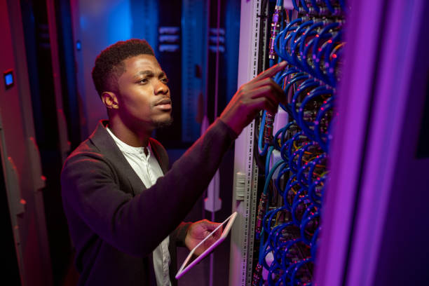 Concentrated Black IT Engineer Checking Connection Concentrated young Black IT engineer standing with tablet in data center and checking connection of cables man and machine stock pictures, royalty-free photos & images