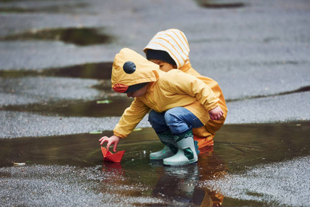 zwei kinder in gelben wasserdichten umhängen und stiefeln, die nach dem regen zusammen mit einem handgefertigten papierboot im freien spielen - puddle rain boot water stock-fotos und bilder