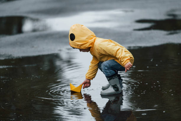 kind in gelbem wasserdichtem umhang und stiefeln, das nach dem regen mit handgefertigtem bootshandy aus papier im freien spielt - puddle rain boot water stock-fotos und bilder