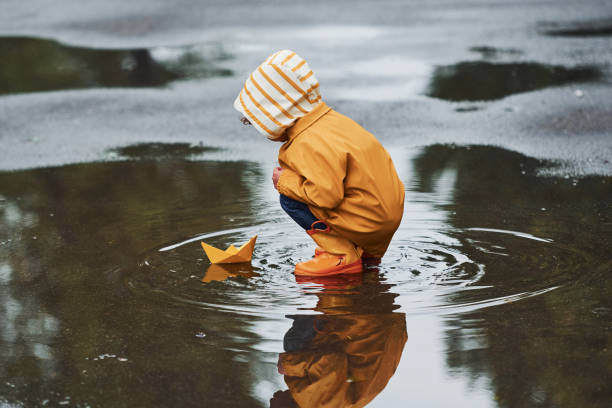 kind in gelbem wasserdichtem umhang und stiefeln, das nach dem regen mit handgefertigtem bootshandy aus papier im freien spielt - puddle rain boot water stock-fotos und bilder