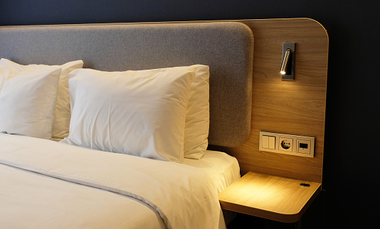 Close-up fragment of bedroom with empty bedside table, reading lamp and USB socket in modern interior​ design home or hotel. Soft pillow and blanket, stylish comfortable furniture.