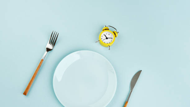 istock Intermittent fasting, diet, lunch time concept. Yellow alarm clock, empty plate and cutlery on a blue background. 1328303792