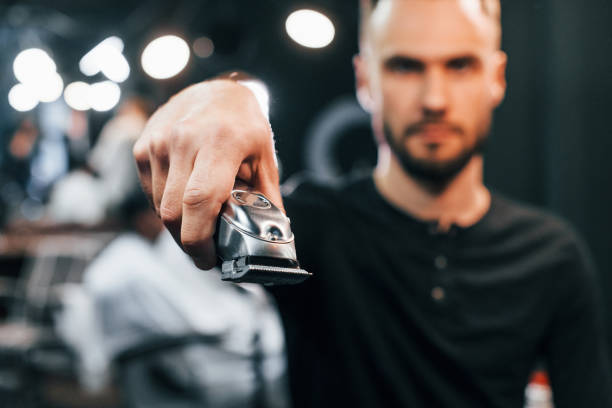 istock Young bearded man standing in barber shop and holding clipper 1328305857