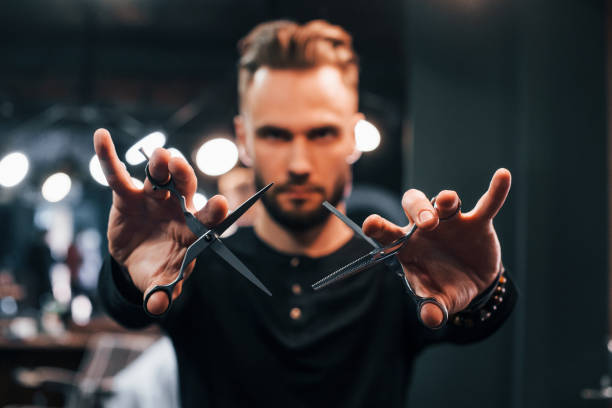 Young bearded man standing in barber shop and holding scissors Young bearded man standing in barber shop and holding scissors. hair stylist stock pictures, royalty-free photos & images