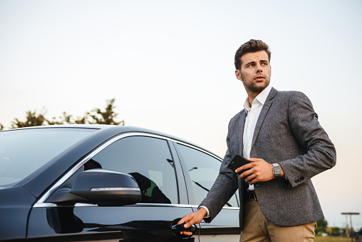 Confident young businessman in suit opens his car door, holding mobile phone