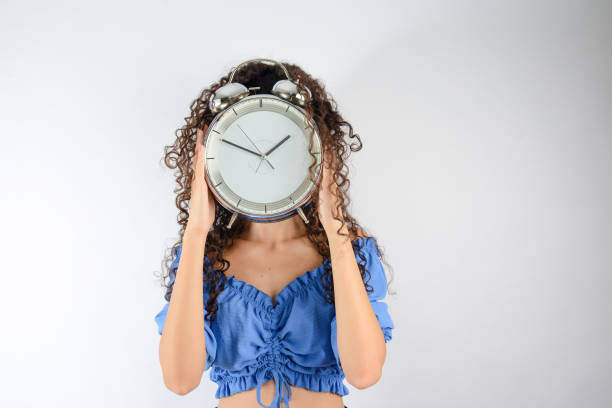 uma garota bonita de cabelo encaracolado está segurando um relógio analógico no rosto. - clock clock face blank isolated - fotografias e filmes do acervo