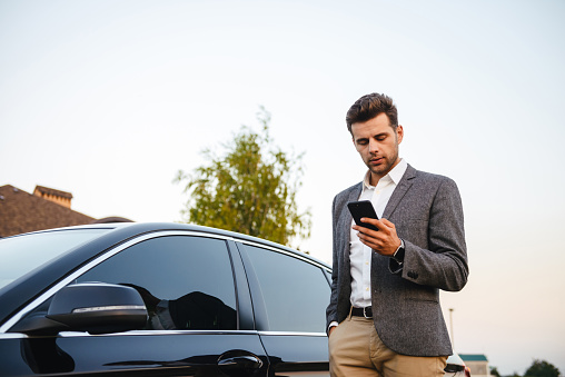 Portrait of rich businessman wearing suit, standing near his luxury black car and using smartphone while holding in hand