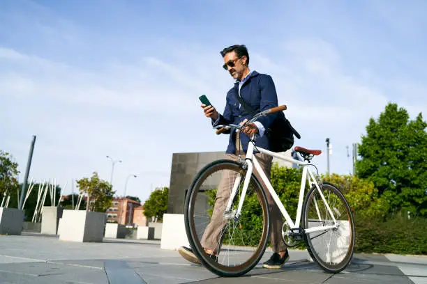Adult male walking to the office with one hand holding the bicycle and the other hand looking at his mobile phone. Eco-friendly transportation concept. High quality photo