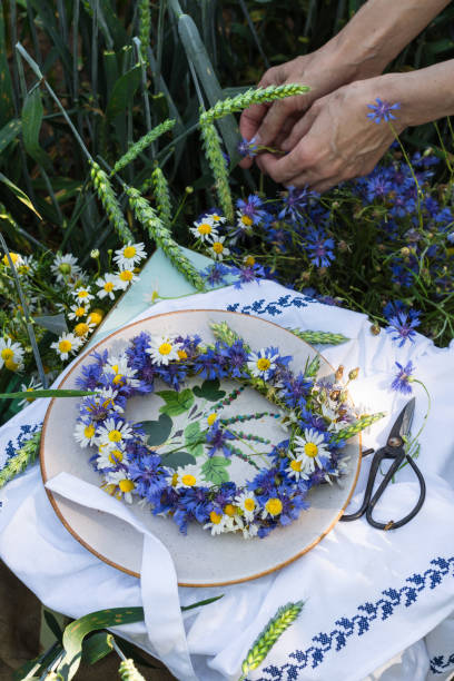 istock Wreath of cornflowers and chamomile on a stool with white tablecloth in wheat field background and hands of woman gathering cornflowers 1328314405