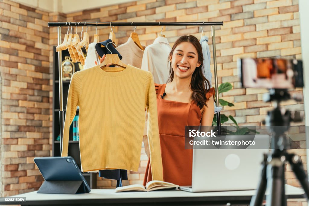 Woman influencer selling clothes online Young and beautiful Asian woman blogger showing clothes in front of smartphone camera while recording vlog video and live streaming at her shop Selling Stock Photo