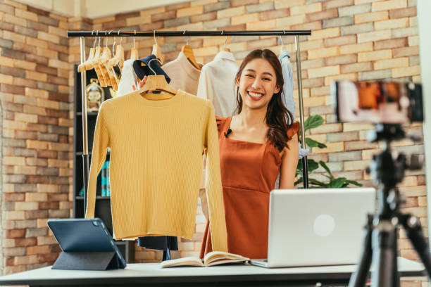 Woman influencer selling clothes online Young and beautiful Asian woman blogger showing clothes in front of smartphone camera while recording vlog video and live streaming at her shop how to sell my photography online stock pictures, royalty-free photos & images