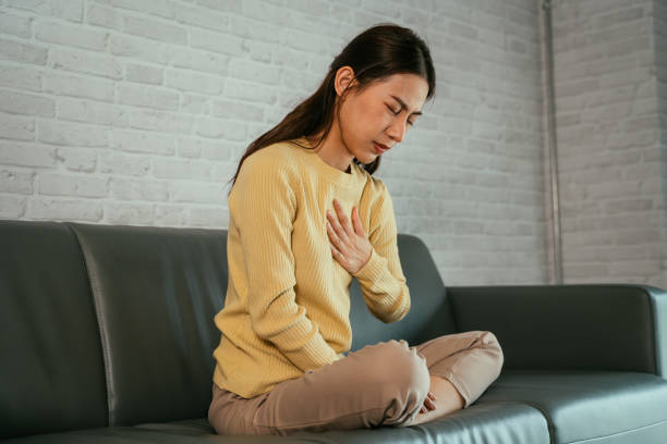 Young female suffering from heartburn Asian young woman feeling discomfort as suffering from heartburn holding chest with closed eyes and sitting with folded legs on couch at home dyspepsia stock pictures, royalty-free photos & images