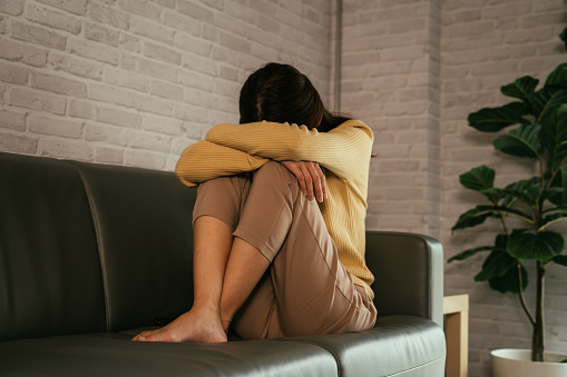 Young woman sitting on couch at home hiding face and hugging knees while feeling depressed and lonely