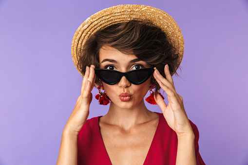 Beauty brunette woman in dress, straw hat and sunglasses posing while looking at the camera over purple background