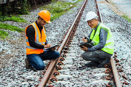 Inspector Engineering wearing helmet and vest worker unifrom checking railway construction work on rail track. Asian workers and coworkers stand beside old train bogies.