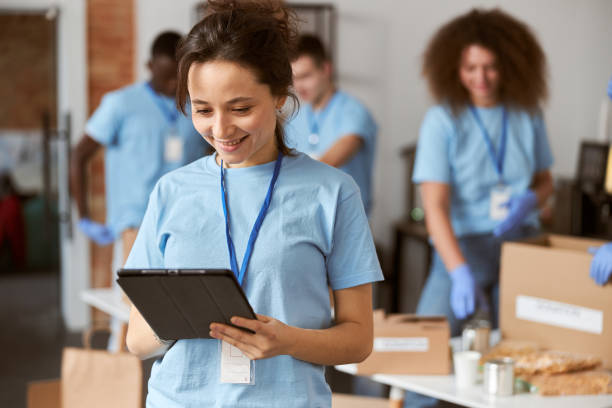 istock Portrait of charming young female volunteer in blue uniform using tablet pc and smiling while standing indoors. Team sorting, packing items in the background 1328325147