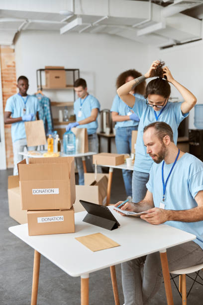 istock Team of diverse volunteers in blue uniform making notes, calculating, sorting and packing items in cardboard boxes while working together on donation project indoors 1328325197