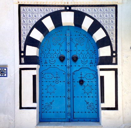 Traditional Tunisian door in Sidi Bou Said with black metal detailing and knobs. Bold, Graphic black and white arch surrounding door and blue and white tiles above.