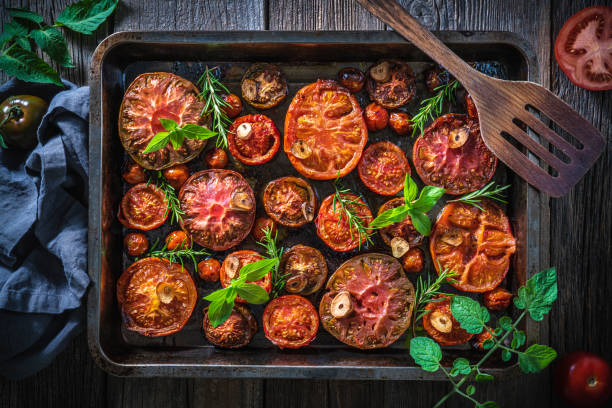 Roasted tomatoes cut varied in baking tray and ladle with basil and rosemary on wood Roasted tomatoes cut varied in baking tray and ladle with basil and rosemary on wood and tomato plant leaves baking sheet stock pictures, royalty-free photos & images