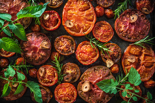 Roasted tomatoes cut varied in baking tray with basil and rosemary macro detail