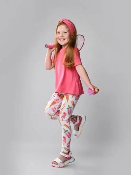 Photo of Happy redhead kid girl in pink t-shirt, colorful pants and sandals is walking going to play with badminton