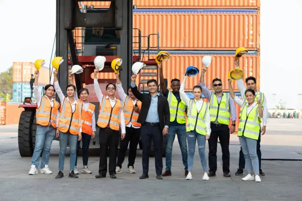 Photo of portrait factory workers or engineers team holding helmet and raise hand up for success in containers warehouse storage
