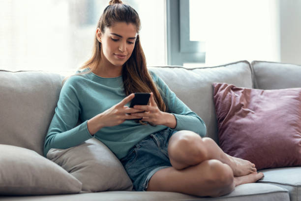 istock Pretty young smiling woman using mobile phone while taking a break sitting on a couch at home. 1328332808