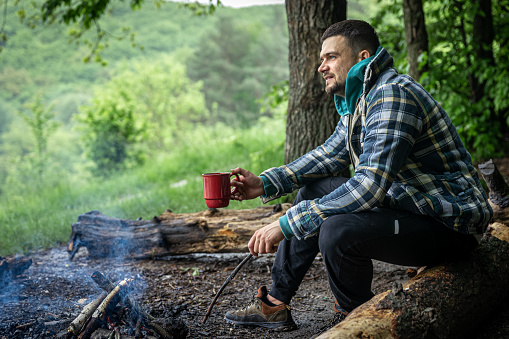 A man with a cup of warming drink warms himself by the fire in the forest.