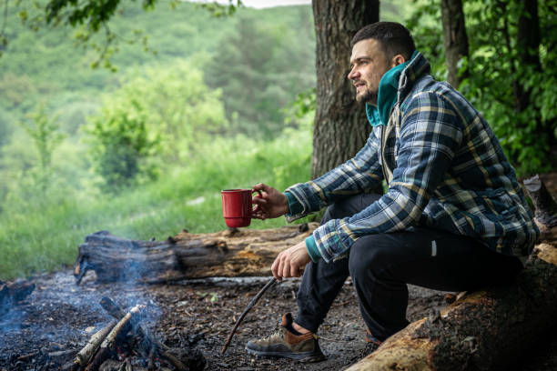 A man on a hike sits by the fire and warms himself. A man with a cup of warming drink warms himself by the fire in the forest. warms stock pictures, royalty-free photos & images