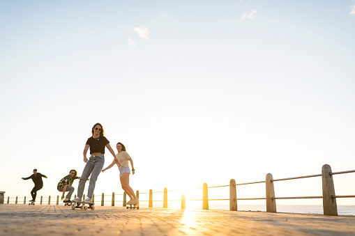 Wide shot group of young adults skateboarding together on the promenade at sunset