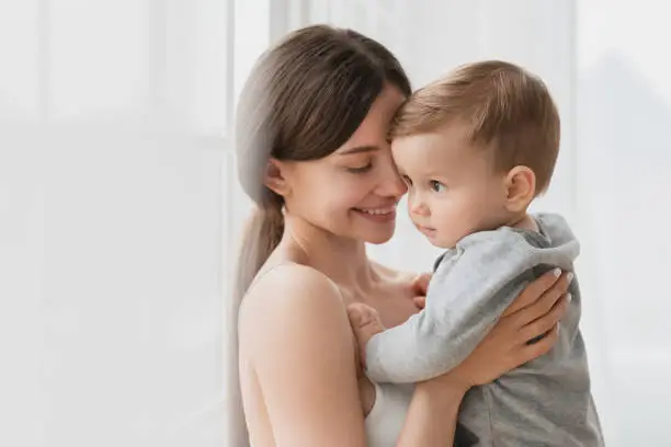Cheerful young single mother hugging embracing little small son daughter baby newborn infant toddler indoors. Motherhood and childcare concept. Postnatal period