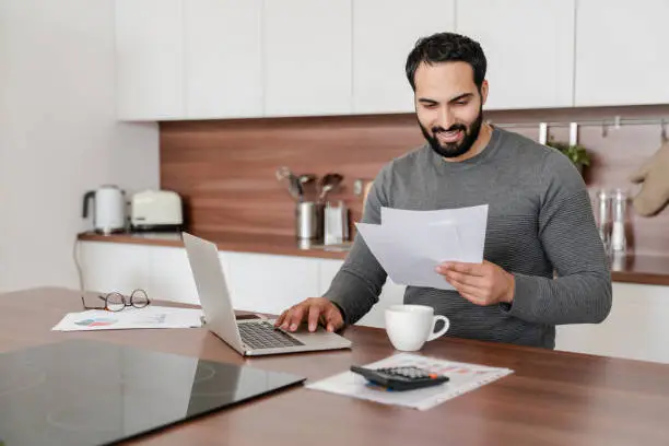 Busy concentrated Middle Eastern man freelancer student counting calculating costs, expenses, income, rental taxes, doing paperwork in the kitchen using laptop. Remote work occupation, finance concept