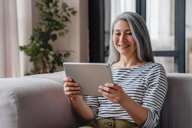 расслабленная смеющаяся зрелая женщина средних лет мама-фрилансер читает электронные книги, использует планшет для просмотра фильмов, веб - grandparent family reading inside of стоковые фото и изображения