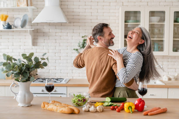 glückliche fröhliche, fröhliche familieneltern mittleren alters, die zusammen in der küche tanzen, essen für ein romantisches abendessen zubereiten, zeit miteinander verbringen. aktive senioren - leben im stock-fotos und bilder