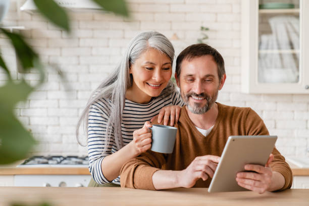 happy mature middle-aged family couple parents husband and wife checking emails, reading news on digital tablet during breakfast, choosing new house, using application online - olhando imagens e fotografias de stock
