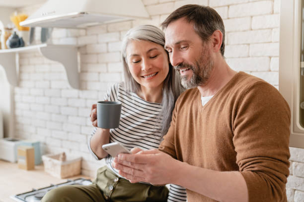 Mature family middle-aged wife and husband couple using smart phone watching videos, social media, reading news on cellphone at home in the kitchen together. Mature family middle-aged wife and husband couple using smart phone watching videos, social media, reading news on cellphone at home in the kitchen together. couple isolated wife husband stock pictures, royalty-free photos & images