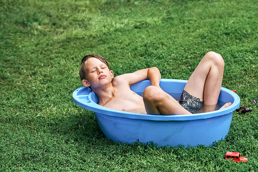 Relaxed preteen boy in shorts lies in large blue plastic basin with water on green lawn grass in shady yard of summerhouse