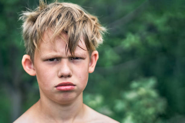 Offended junior schoolboy with blond rough hair pouts posing for camera in green park on nice summer day close view