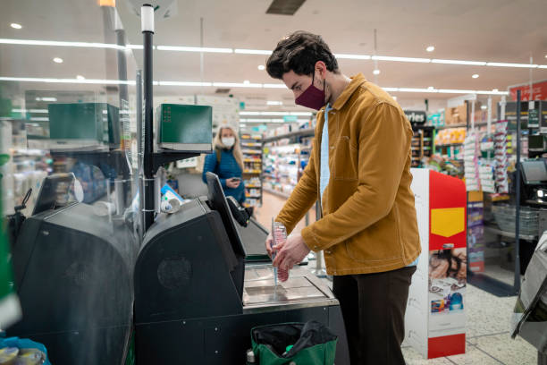 Using Self Service Till At Supermarket A side view of a young man scanning his items at the self service checkout at the supermarket in Whitley Bay in the north-east of England he is wearing a protective face covering to protect himself and others during the ongoing pandemic. self checkout stock pictures, royalty-free photos & images